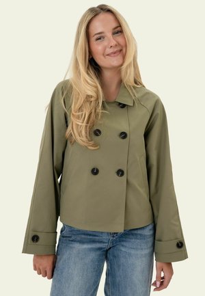 Młoda kobieta z długimi blond włosami, ubrana w oliwkowozielony, dwurzędowy płaszcz i niebieskie dżinsy, lekko się uśmiecha.