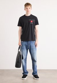 Zwarte katoenen t-shirt met een rood en wit logo, gecombineerd met blauwe spijkerbroek en zwarte sneakers, met een zwarte totebag in de hand.
