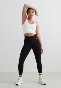 Kvinde i hvid crop top, sorte højtaljede leggings, stribede sokker og hvide sneakers, poserer med en arm løftet mod en ensfarvet væg.