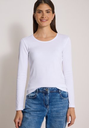 Jeune femme aux longs cheveux foncés portant une chemise blanche à manches longues et un jean bleu, souriant à la caméra sur un fond clair.