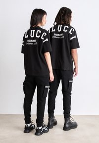 Zwarte oversized t-shirts met de tekst "LUCCI" en "Established Milano 1978" gedrukt op de achterkant, gecombineerd met zwarte cargobroeken en laarzen.