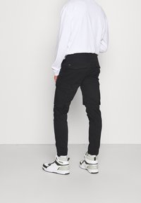 Zwarte cargobroek met meerdere zijzakken, slim fit en stevig materiaal. Gekenmerkt door het dragen van hoge witte en groene sneakers.