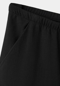 Gros plan sur un pantalon en tissu noir avec une taille élastique et des détails de couture latérale sur un fond clair.