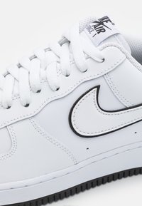 Fehér Nike Air Force 1 sneaker, sima bőrből készült, klasszikus sziluettel, kerek orral, fehér fűzőkkel és fekete akcentusú swoosh logóval.