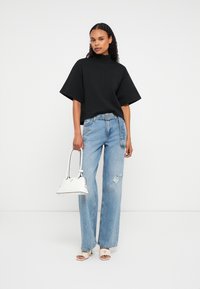 Frau mit langen lockigen Haaren, die ein schwarzes oversized Oberteil, helle blaue, gefragte Jeans und weiße High Heels trägt und eine weiße Handtasche hält.