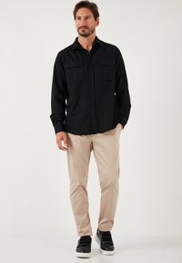 Cămășă neagră cu mâneci lungi și nasturi, având două buzunare la piept, purtată cu pantaloni bej și adidași negri, pe un fundal neutru.