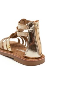 Metaalachtige gouden sandal met uitsnijdingen, verstelbare banden en een zijrits. Voorzien van een getextureerde tan inlegzool en een bruine rubberen zool.
