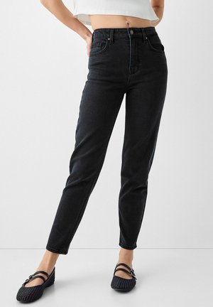 Frau trägt schwarze High-Waist-Jeans, weißes kurzes Oberteil und schwarze geflochtene Ballerinas vor einem schlichten weißen Hintergrund.