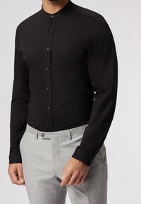 Camisa negra de manga larga con cuello mao, que presenta una tapeta delantera con botones negros, combinada con pantalones grises claros a medida.