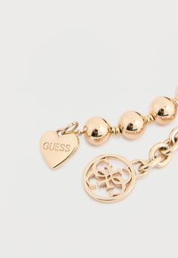 Guldtonet armbånd med glatte, runde perler, et hjerteformet charm med indgraveringen "GUESS" og en cirkulær logo vedhæng.