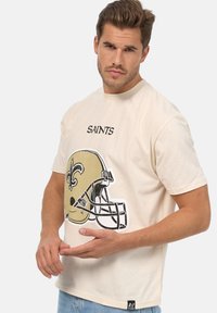 Camiseta beige con un gráfico de un casco de fútbol americano y el nombre del equipo "SAINTS" impreso arriba. Mangas cortas, cuello redondo y corte casual.