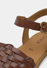 Καφέ δερμάτινο σανδάλι με πλεκτό πάνω μέρος, επίπεδη σόλα και λουράκι με αγκράφα. Στο εσωτερικό εμφανίζεται η ένδειξη μεγέθους 4½. Λείο υλικό με ελάχιστες λεπτομέρειες.