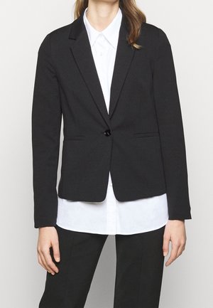 Blazer nero con chiusura a un bottone, rever a scosse e due tasche frontali, indossato sopra una camicia bianca. Tessuto liscio e vestibilità su misura.