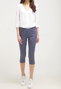 Szare spodnie 3/4 o slim fit z zapięciem na guzik z przodu i kieszeniami po bokach, w połączeniu z białą bluzką z długim rękawem.