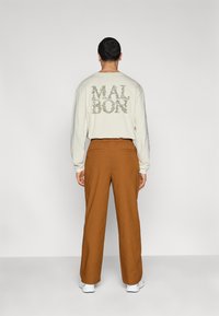 Chemise crème à manches longues avec un graphisme "MALBON" en vert dans le dos, associée à un pantalon large marron et des baskets blanches.