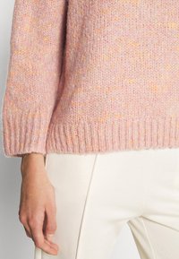 Rosa stickad tröja med en texturerad yta, med ribbat avslut och nederkant, kombinerad med vita byxor för en kontrasterande look.