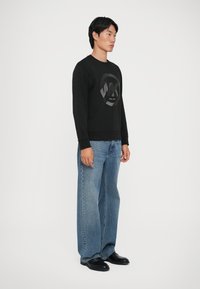 Μαύρη φόρμα με κυκλικό σχέδιο λογότυπου, συνδυασμένη με ανοιχτό μπλε φαρδιά τζιν παντελόνια και μαύρα παπούτσια. Λευκό απλό φόντο.