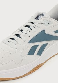 Λευκό και μπλε αθλητικό παπούτσι Reebok με κορδόνια, υφασμάτινο άνω μέρος από σουέτ με υφή και σόλα από καουτσούκ, εμφανίζεται σε κοντινό πλάνο από το πλάι.