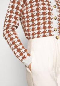 Cardigan à manches longues à motif pied-de-poule marron et blanc avec des boutons beige, associé à un pantalon crème taille haute, main dans la poche.