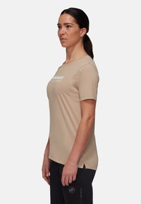 Beige t-shirt med korte ærmer, der har et hvidt logo på brystet, rund halsudskæring og subtile sideslidser. Fremstillet af blødt, glat stof.