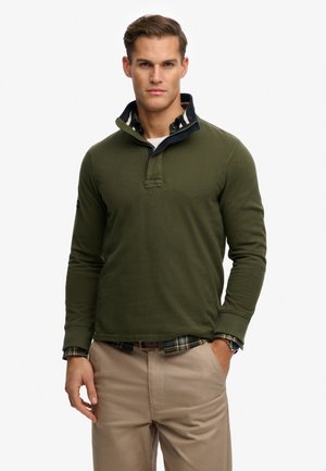 Młody mężczyzna noszący oliwkowy sweter na koszuli w kratę, beżowe spodnie, z jedną ręką w kieszeni na jednolitym białym tle.