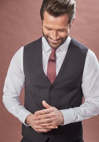 Vesta gri închis peste o cămașă albastră deschisă, asociată cu o cravată roșie texturată. Bărbatul își ajustează mâinile, zâmbind ușor.