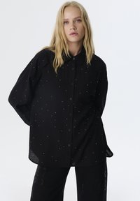 Camicia nera con bottoni, caratterizzata da un motivo testurizzato e decorata con punti bianchi distribuiti in tutto il tessuto. Maniche lunghe e vestibilità comoda.