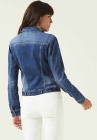 Donkerblauwe denimjack met een getailleerd ontwerp, voorzien van lange mouwen, een knoopsluiting aan de onderkant en lichte stikseldetails langs de naden.