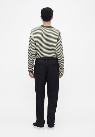 Homme debout de dos, portant une chemise à manches longues rayée verte et blanche, un pantalon noir et des chaussures noires avec des semelles blanches.