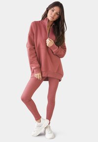 Dammig rosa zip-up sweatshirt med hög krage och ribbat fåll, matchad med leggings. Modellen bär vita sneakers. Slät textur.