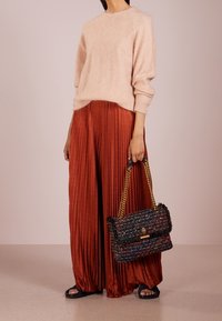 Žena v světle růžovém svetru a rezavě oranžových širokých kalhotách s plisem, držící černou kabelku z tkaniny s zlatými řetízkovými popruhy.