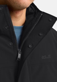 Veste noire en tissu soft-shell avec col haut, fermeture éclair à l'avant, boutons-pression et logo. Présente une texture lisse et des lignes épurées.