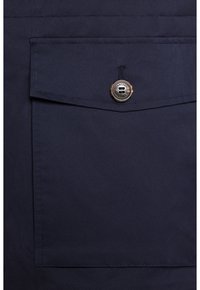 Granatowa kurtka z tkaniny z dużą przednią kieszenią, zapięcie na złotą guziki oraz minimalistyczne detale przeszycia. Gładka faktura, nowoczesny design.