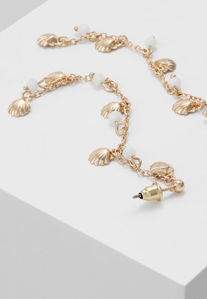 Bracelet chaîne en or avec petites perles blanches et breloques coquillages dorées sur une surface blanche.
