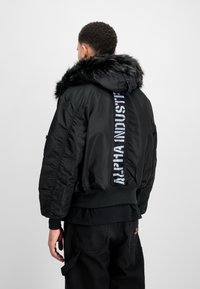 Schwarze Bomberjacke mit Kunstfellkapuze, lockere Passform, mit einem vertikalen weißen Logo-Print auf dem Rücken sowie gerippten Bündchen und Saum.