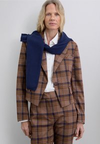 Brown and blue plaid suit with a cropped jacket and fitted trousers, layered with a navy blue sweater casually tied around the shoulders.