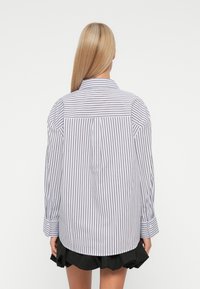 Persoon met lang blond haar draagt een oversized wit shirt met verticale zwarte strepen en een zwarte gerimpelde rok, gezien van achteren.