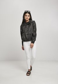 Must värvi kerge anorak, millel on esipocket, reguleeritav kapuuts ja elastsed varrukad, kantud musta topi ja valgete teksade üle, koos mustade sandaalidega.