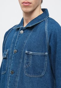Kurtka dżinsowa w ciemnym niebieskim kolorze z ostrym kołnierzem, srebrnymi zatrzaskami, dwoma przednimi kieszeniami oraz widocznymi detalami szwów.
