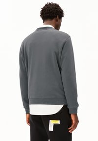 Grijze sweatshirt met een kraag en geribbelde manchetten, met een gelaagd ontwerp over een wit shirt en een label op de achterkant.