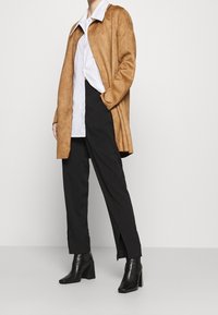 Brun mocka lång kappa över en vit skjorta, tillsammans med svarta byxor och svarta klackstövlar. Kappan har krage och sidofickor.