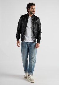Schwarze Leder-Bomberjacke mit Reißverschluss, zwei Taschen, gerippten Bündchen, getragen über einem weißen T-Shirt und kombiniert mit hellblauen Jeans und Sneakers.