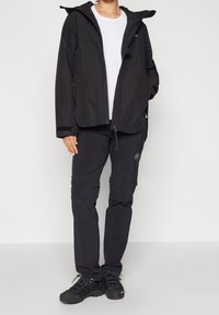 Personne portant une veste d'extérieur noire, un pantalon noir avec un logo sur la cuisse, une chemise blanche et des chaussures de randonnée noires, debout avec une main dans la poche.