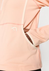 Ροζ ανοιχτόχρωμο μακρυμάνικο τοπ κατασκευασμένο από υφασμάτινη υφή, με φερμουάρ στο λαιμό, λεπτομέρειες σε κρέμ χρώμα και πλαϊνή τσέπη με κεντημένο λογότυπο.