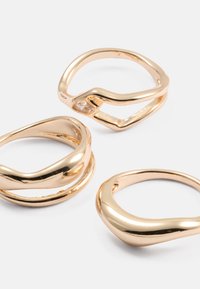 Tres anillos de oro con diseños suaves y ondulados; uno presenta una pequeña piedra preciosa transparente. Cada anillo exhibe un acabado pulido y formas distintas.