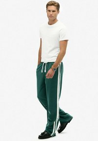 Groene joggingsbroek met witte zijstrepen, met een trekkoord in de taille, gecombineerd met een eenvoudig wit T-shirt en zwarte schoenen. Het model staat in profiel.