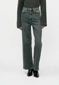 Gai zaļi brīva piegriezuma džinsi, kas izgatavoti no denima, ar augstu jostasvietu, vieglu izbalējumu un kontrastējošu šuvumu, apvienoti ar melnām papēžu kurpēm.