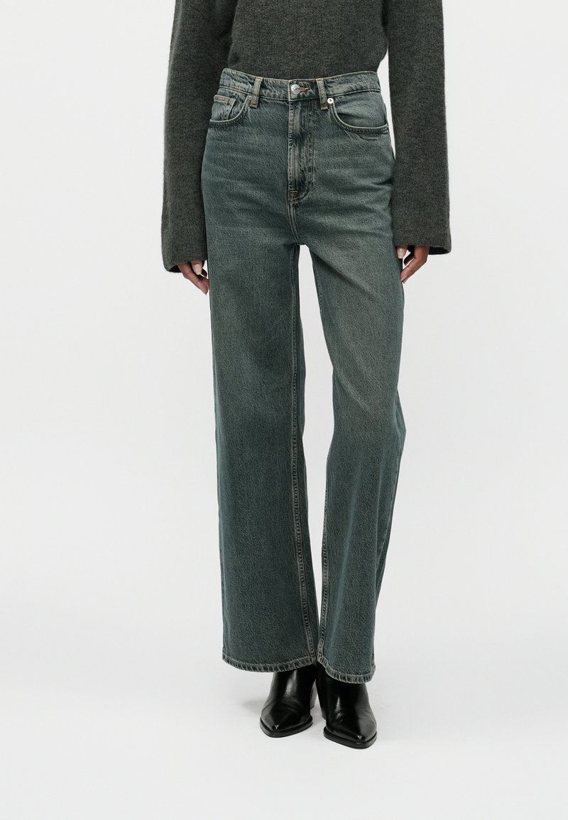 Gai zaļi brīva piegriezuma džinsi, kas izgatavoti no denima, ar augstu jostasvietu, vieglu izbalējumu un kontrastējošu šuvumu, apvienoti ar melnām papēžu kurpēm.