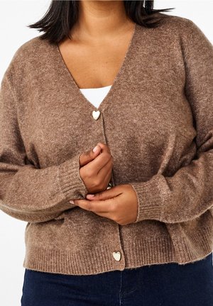Cardigan marron tricoté avec des boutons en forme de cœur, porté sur un haut blanc, les mains doucement croisées à la taille.