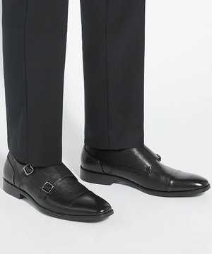 Chaussures habillées en cuir noir à double boucle portées avec un pantalon formel noir sur un fond clair uni.